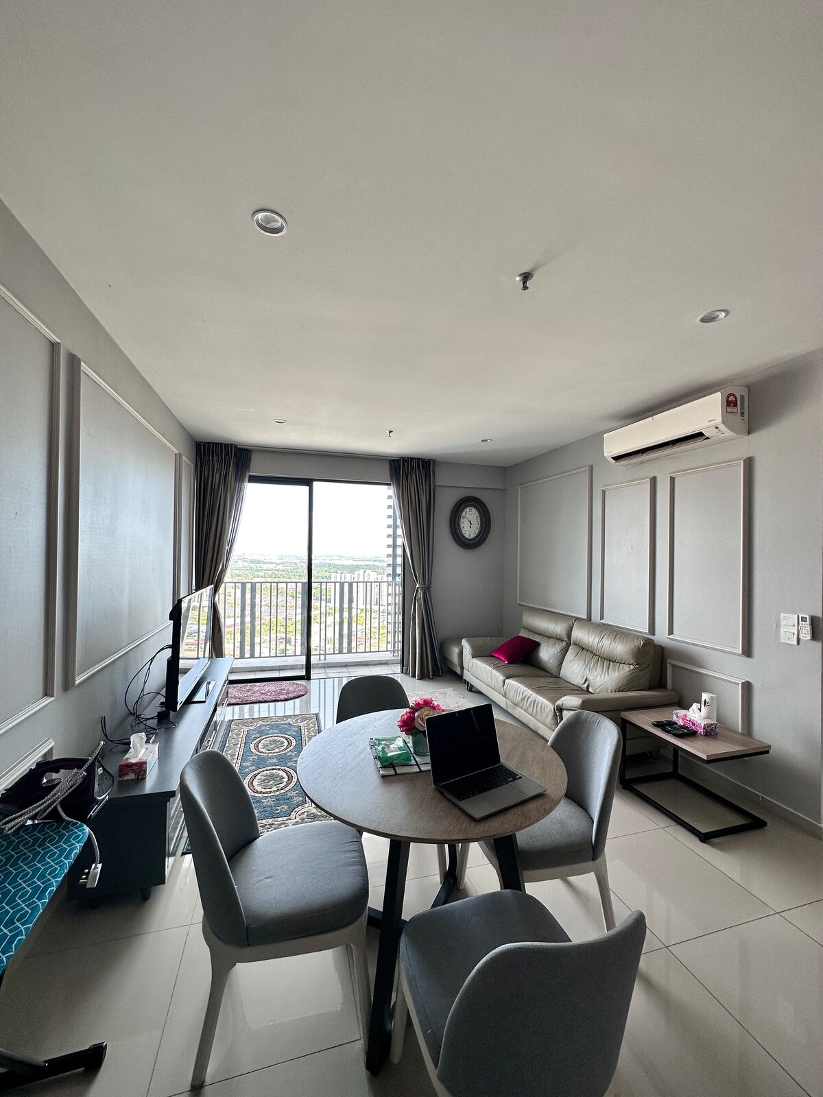 The living area features a comfortable sofa positioned near a balcony with a skyline view. A dining table with four chairs is placed centrally, accompanied by a television on a console. Light-colored walls and large windows create an airy atmosphere.