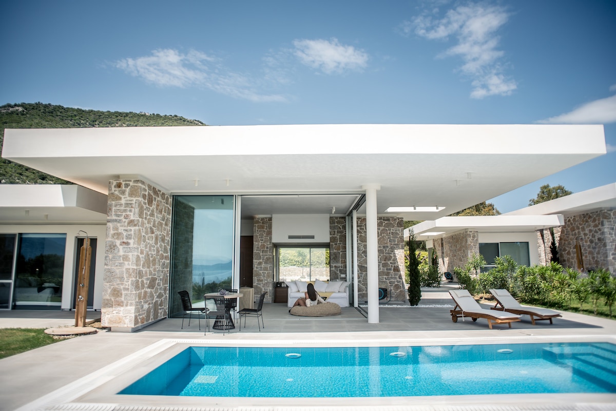 A modern villa is shown with sleek architecture, featuring a covered outdoor seating area beside a clear blue pool. Stone walls contrast with large glass elements, while sun loungers are placed around the pool, surrounded by lush greenery and mountainous views.