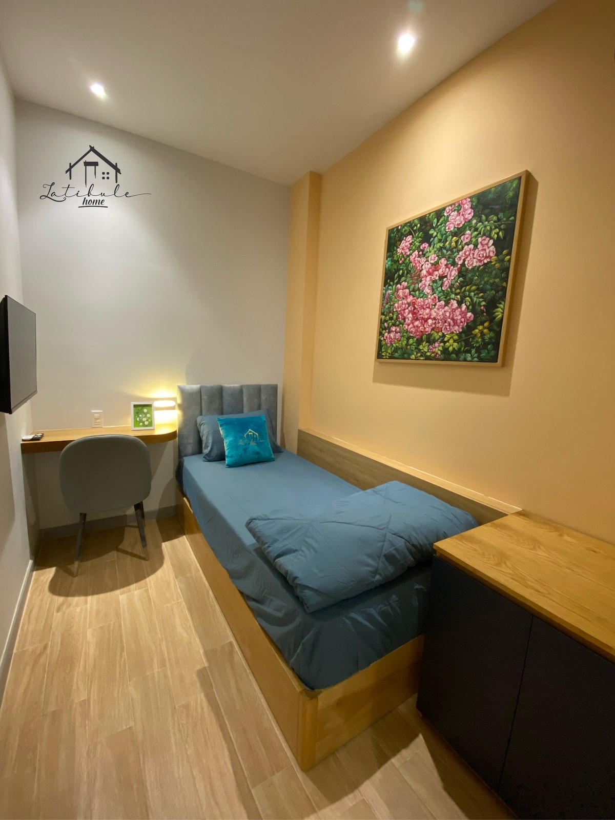 A compact room designed for a solo traveler features a single bed with muted blue bedding. A small desk and chair are positioned next to the bed, with a wall-mounted television nearby. A vibrant floral painting adorns the wall, adding a touch of color to the minimalist decor.