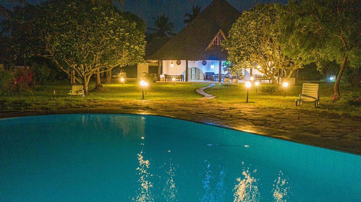 A tranquil evening scene showcases a swimming pool reflecting soft lights. Lush gardens with scattered trees frame the area, while a spacious cottage is illuminated in the background, featuring an inviting entrance and outdoor seating.