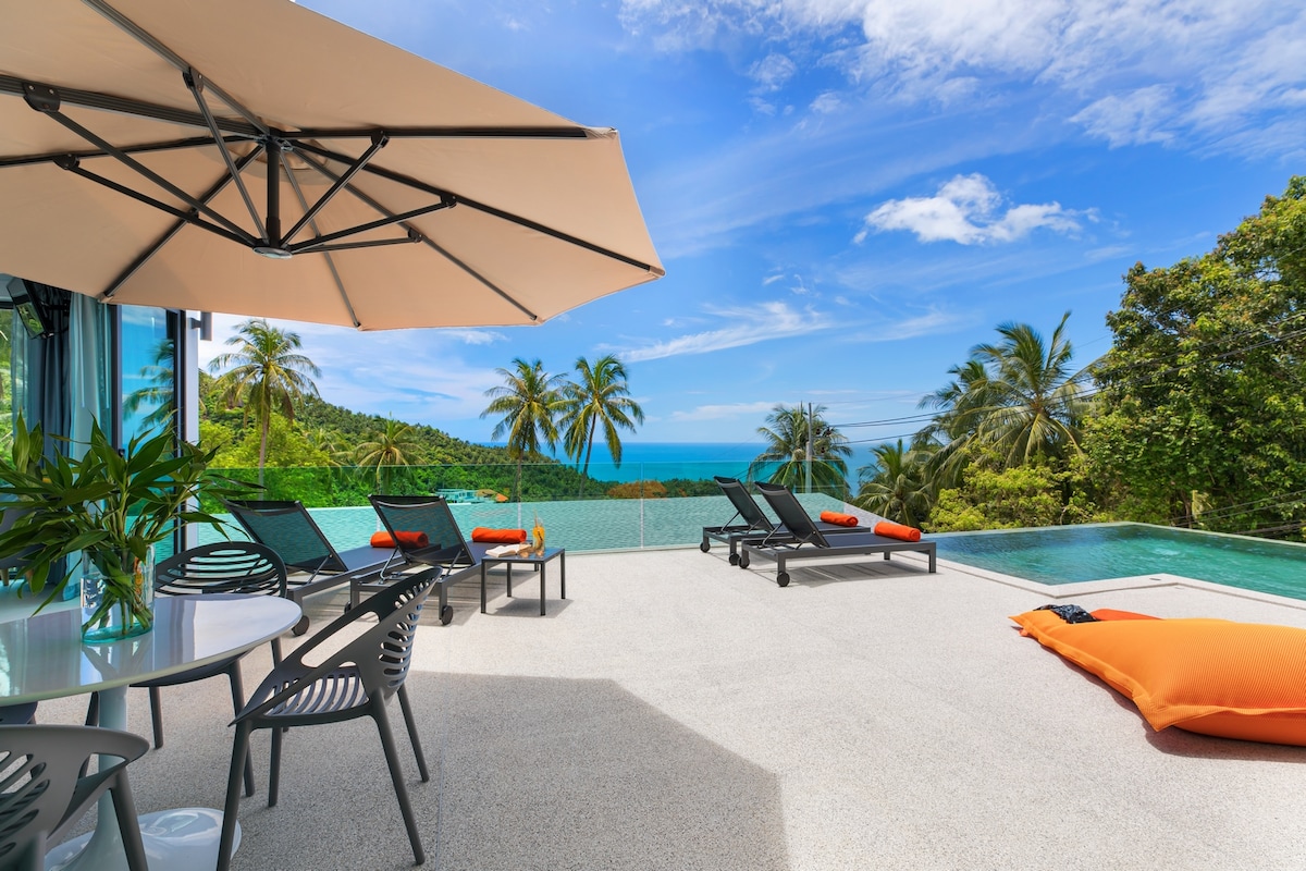 A sun-drenched terrace features a salt-water infinity pool overlooking a vast jungle landscape and the ocean. Sun loungers with orange cushions are positioned near an outdoor dining area shaded by an umbrella, creating a perfect spot for relaxation.