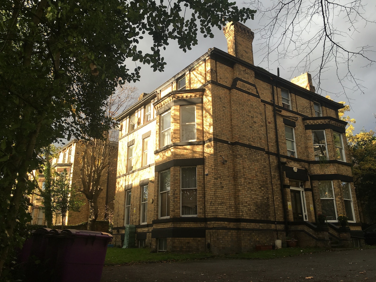 An old brick building with character, featuring tall windows and intricate detailing. A blend of natural and artificial light casts shadows across the façade, highlighting the architectural features. Surrounding trees add greenery to the environment, enhancing the serene atmosphere of the location.