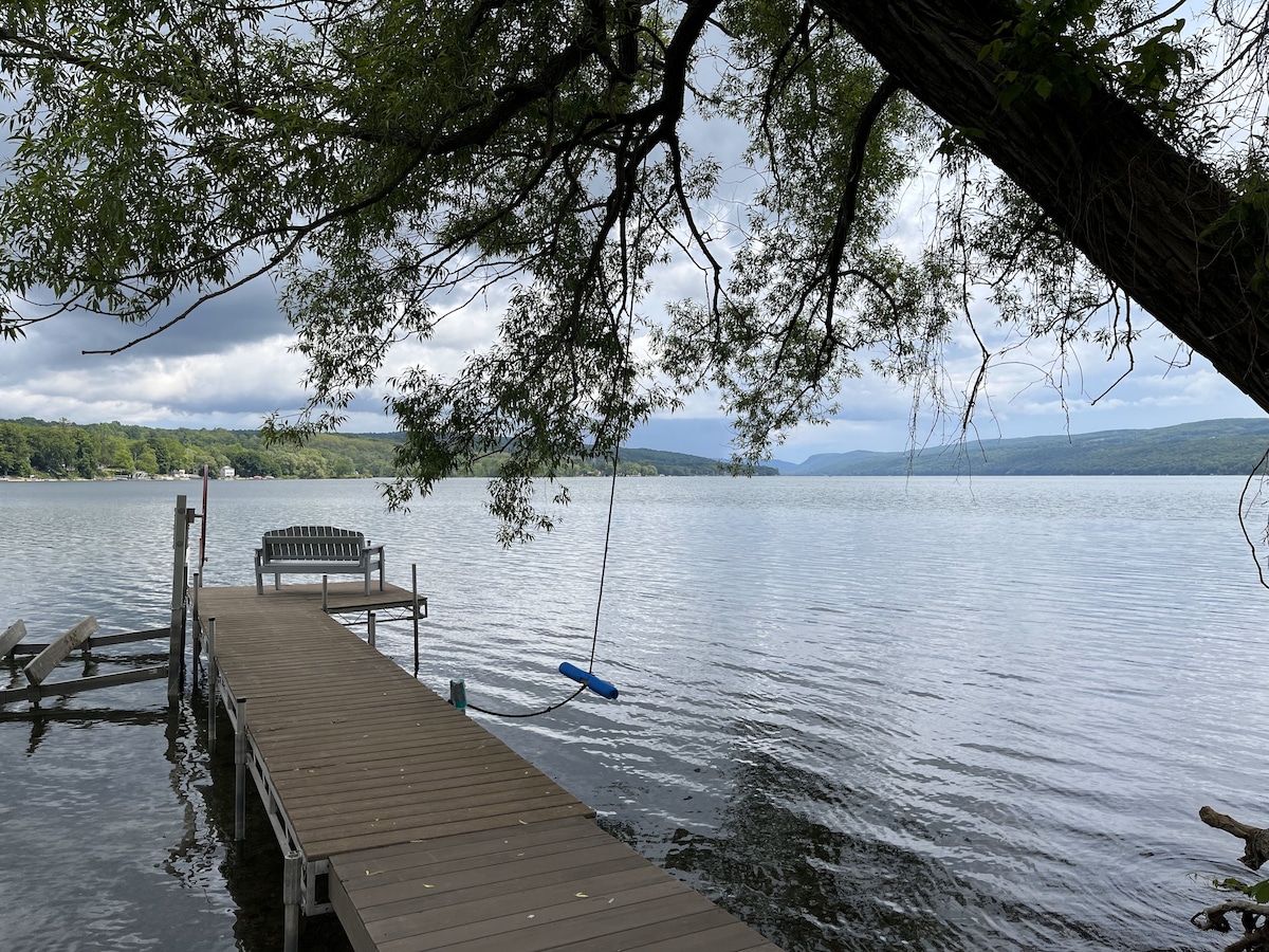 A wooden dock extends over the calm waters of Otisco Lake, framed by lush greenery from overhanging trees. A swing hangs nearby, inviting relaxation by the water's edge, while distant hills subtly rise against the cloudy sky.