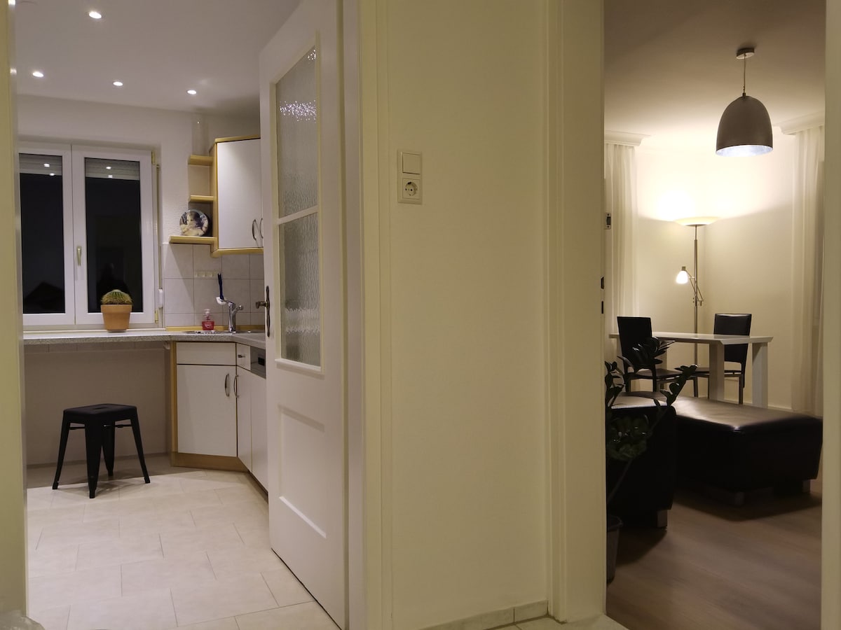 A view of an open kitchen area features light cabinets and a small potted plant atop the counter. The adjoining living area is glimpsed through a doorway, showcasing a dining table with four chairs and a cozy seating arrangement illuminated by overhead lighting.