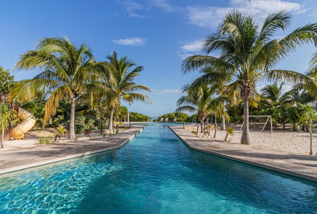 A serene view of a swimming pool lined with palm trees, leading toward a sandy area. The inviting turquoise water glistens under the bright sky, creating a refreshing ambiance ideal for relaxation.