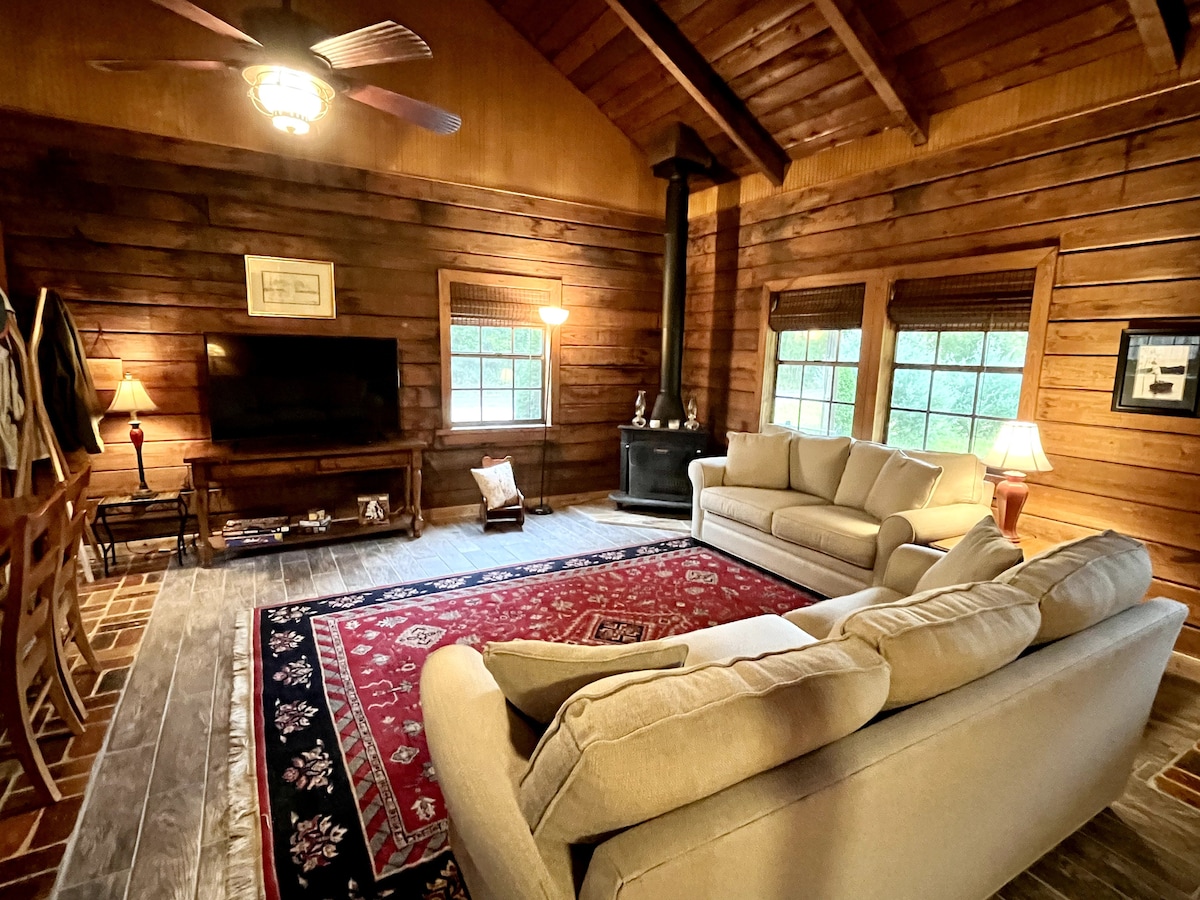 A cozy living area features two comfortable sofas facing a large television. Warm wood paneling covers the walls, and a decorative rug lies on the floor. Natural light filters through two windows, with a wood-burning stove adding to the cabin's inviting ambiance.