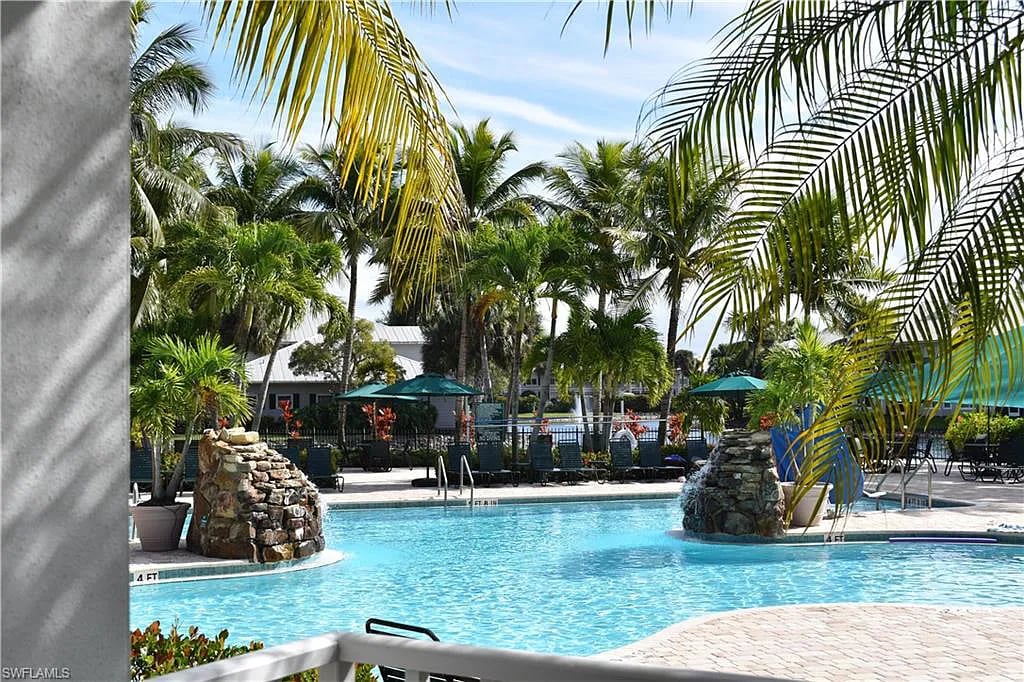The image captures a resort-style pool surrounded by tropical palms and colorful lounge umbrellas. A rock feature creates a central focus in the shallow area of the pool, which is lined with paved walkways, inviting relaxation in a serene environment.