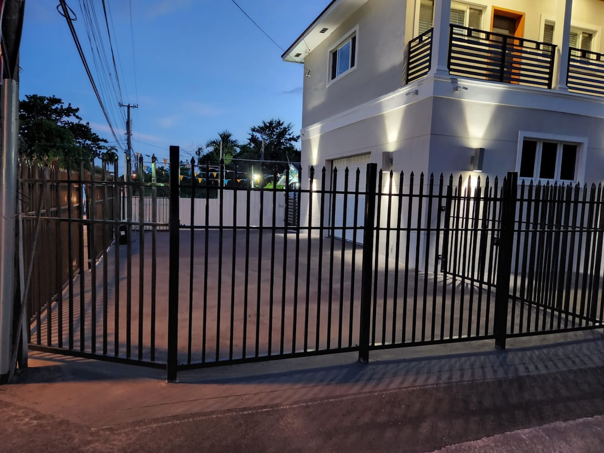 A spacious outdoor parking area is enclosed by a black metal fence, with ample room for multiple vehicles. Soft lighting highlights the structure during twilight, showcasing the clean lines of the modern building behind.