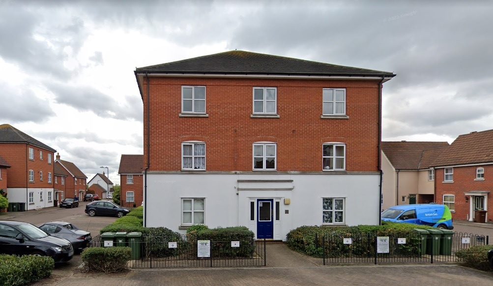 A modern three-story brick building features a symmetrical facade with multiple windows on the upper levels. The entrance is marked by a central white panel below the blue door. Surrounding greenery adds a touch of nature to the structured environment.