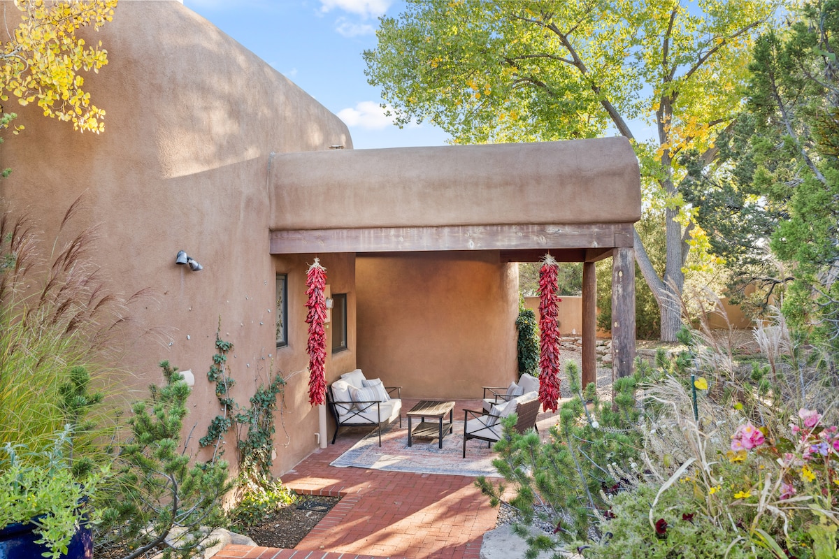 A welcoming outdoor patio area features a rustic adobe structure, with shaded seating arranged on a brick floor. Decorative red chile ristras hang from the roof, complementing the surrounding native plants and trees. Sunlight filters through the foliage, creating a serene ambiance.