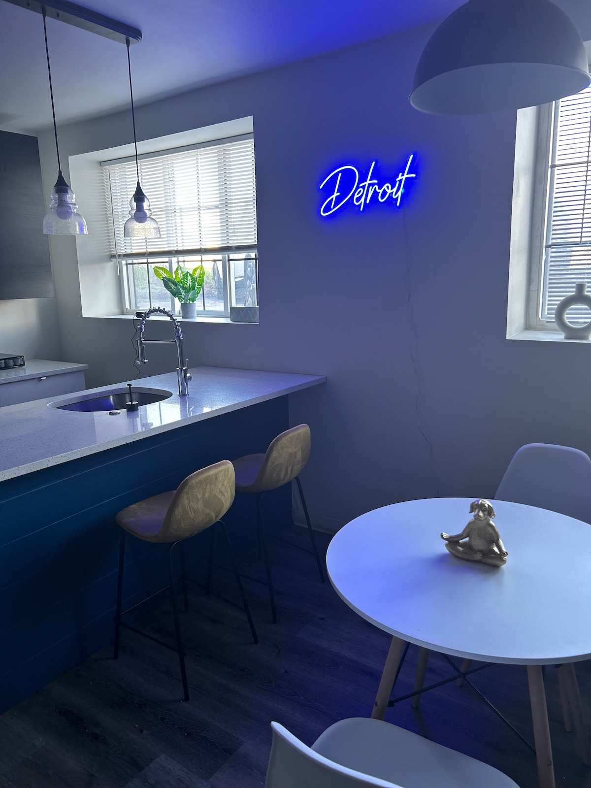 A well-lit kitchen area features a modern design with a central island and bar stools. Large windows allow natural light to illuminate the space, while a neon sign displaying "Detroit" adds character. A small dining table is positioned nearby, enhancing dining options.
