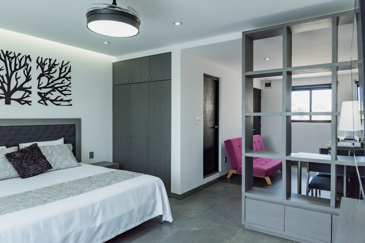 A spacious bedroom features a comfortable bed with decorative linens. A modern grey wardrobe and a glass partition separate a sitting area with a pink lounge chair. Natural light is visible through a large window, enhancing the clean lines of the contemporary design.