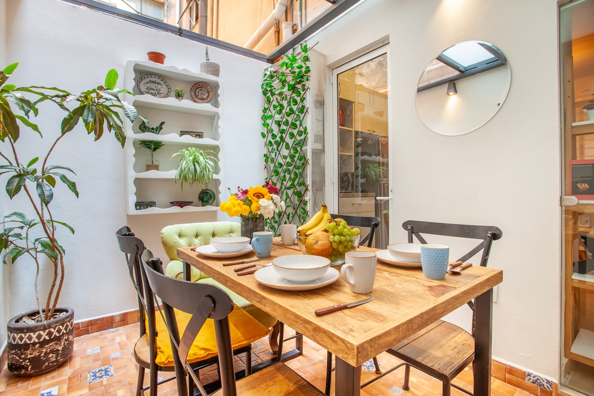 A bright dining area features a wooden table set for meals, surrounded by comfortable chairs. A variety of plants are displayed nearby, contributing to a fresh ambiance. A glass door invites natural light in, while a decorative shelf adds a touch of charm.