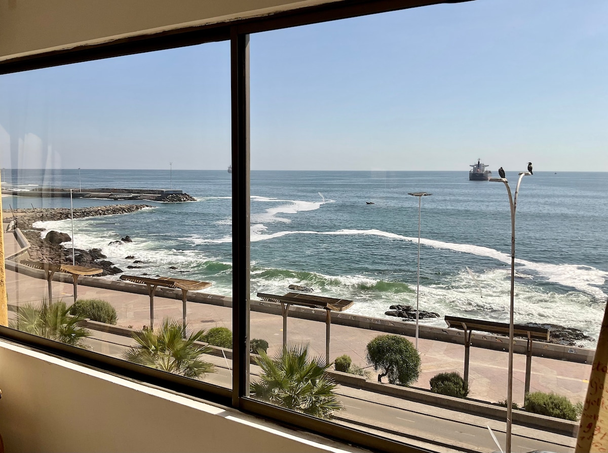 A large window offers a view of the coastline, where waves gently break against the rocky shore. A shimmering blue sea stretches into the distance, and a cargo ship is seen sailing in the horizon. Palm trees line the promenade below, enhancing the scenic waterfront.