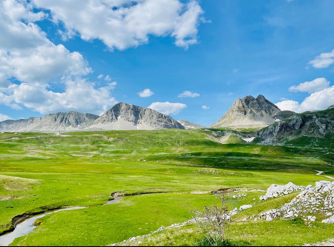 A sweeping view of lush green hills stretches under a bright blue sky, dotted with fluffy white clouds. Majestic mountains rise in the background, showcasing rugged peaks and rocky terrain. A gentle stream winds through the grassy landscape, merging with the natural beauty of the area.