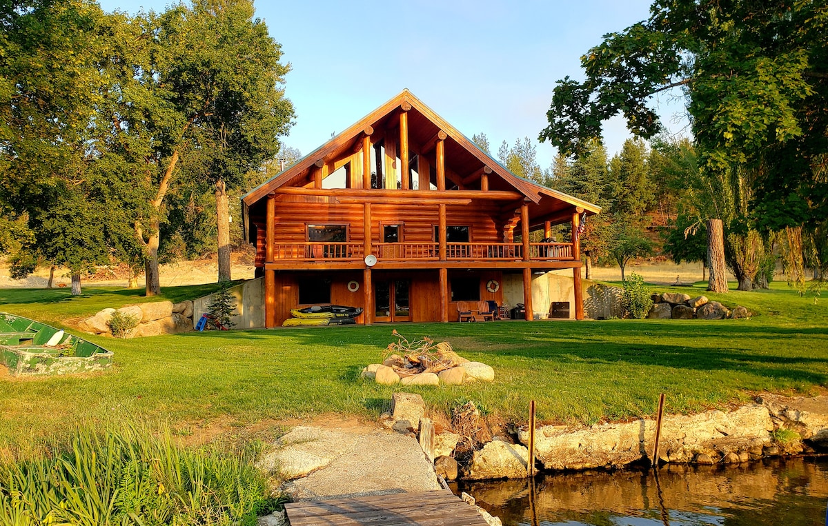 A log cabin-style home is set against a backdrop of lush greenery. The large structure features an expansive porch with wooden railings, highlighting an inviting outdoor space. A stone path leads to the tranquil lakefront, bordered by manicured grass and scattered rocks.
