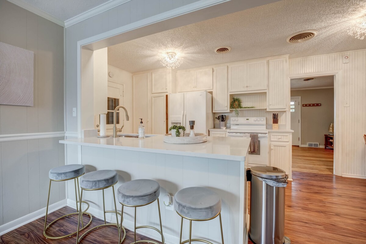 An open-concept kitchen reveals a spacious bar with four upholstered stools. Light wood cabinetry and a white countertop provide a clean aesthetic. The room is brightened by overhead lighting and offers a view into the adjacent living area, enhancing connectivity and comfort.