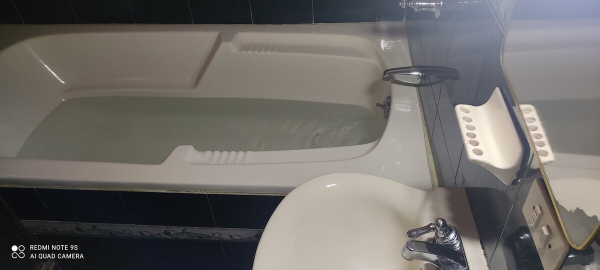 A white bathtub filled with water is positioned alongside a clean sink. The bathroom features dark tiled walls and a large mirror, reflecting the fixtures and water. A small shelf is visible next to the sink, adding functionality to the space.