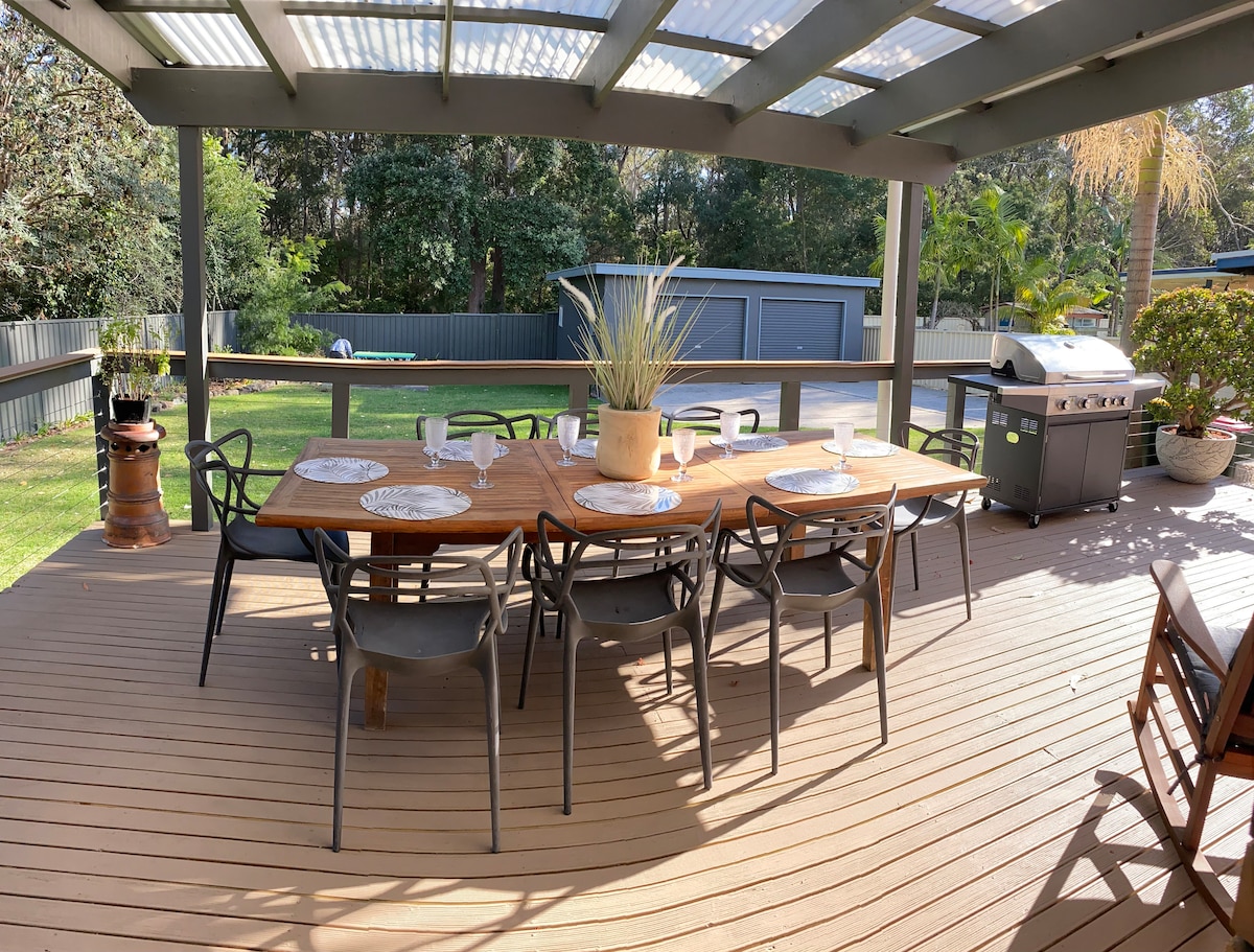 An outdoor dining area is positioned under a covered deck, featuring a large wooden table surrounded by eight chairs. A barbecue grill stands nearby, with lush greenery and a spacious yard visible in the background. Natural light filters through the roof panels.