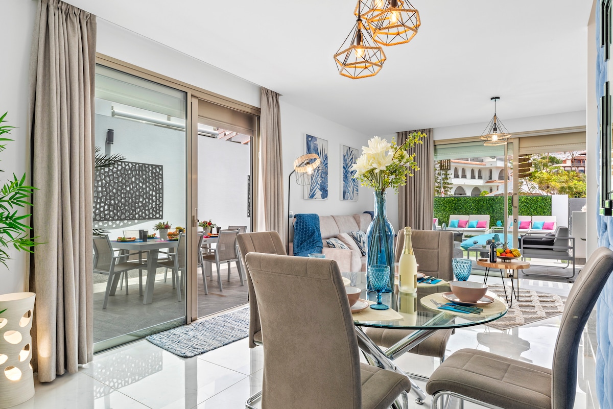 A modern dining area features a sleek glass table surrounded by comfortable chairs. Natural light flows through large sliding doors, revealing an outdoor seating area. Decorative elements include fresh flowers and tasteful tableware, while soft lighting fixtures add a contemporary touch.