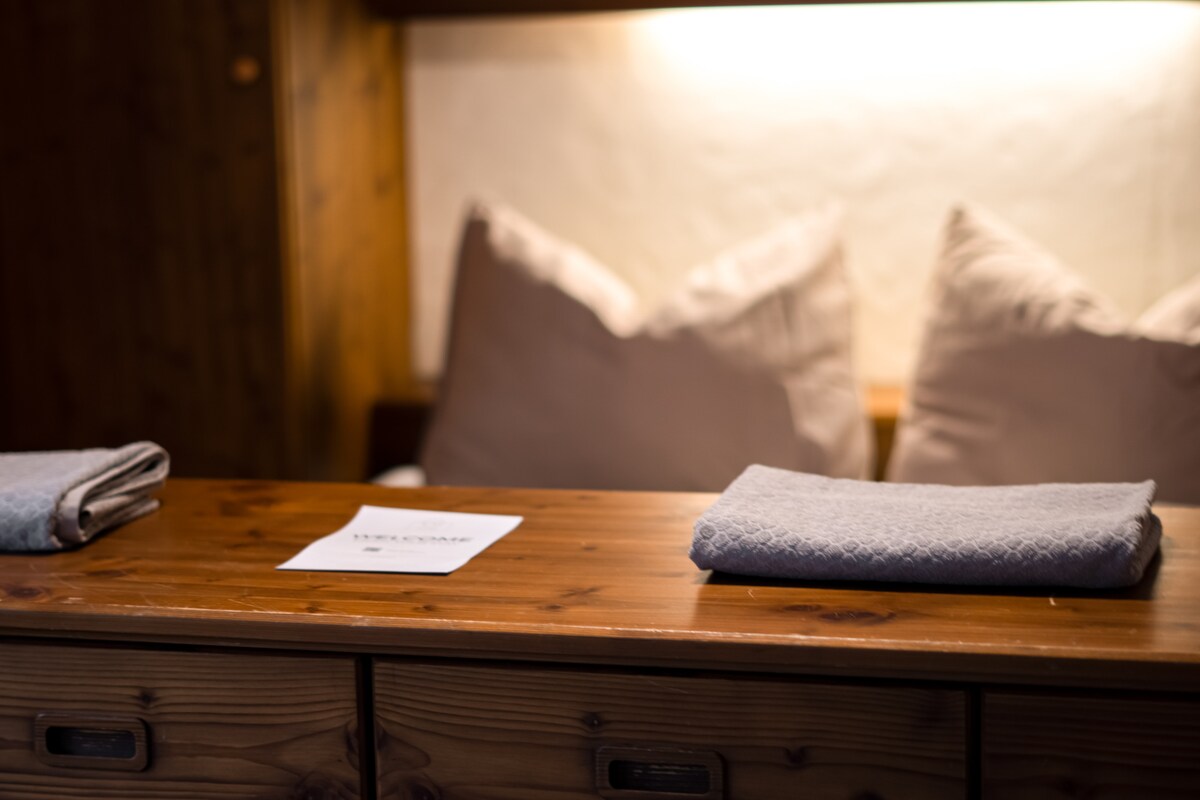 A wooden table is positioned in a cozy corner, featuring neatly folded gray towels. In the background, two soft, plush pillows are arranged against a warm, softly lit wall, contributing to a welcoming and comfortable environment.