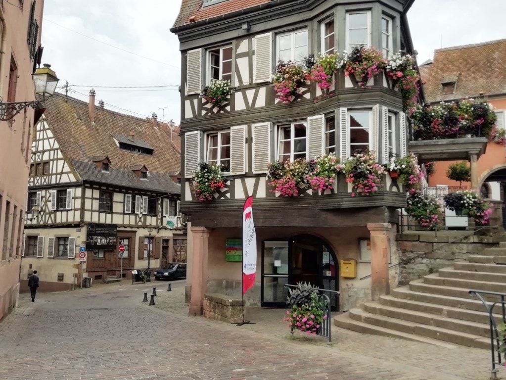 A charming Alsace-style building features traditional timber framing and is adorned with vibrant flower boxes. The cobbled street in front allows for easy pedestrian access. Nearby, another historic structure with a similar architectural style is visible, contributing to the quaint atmosphere of the area.