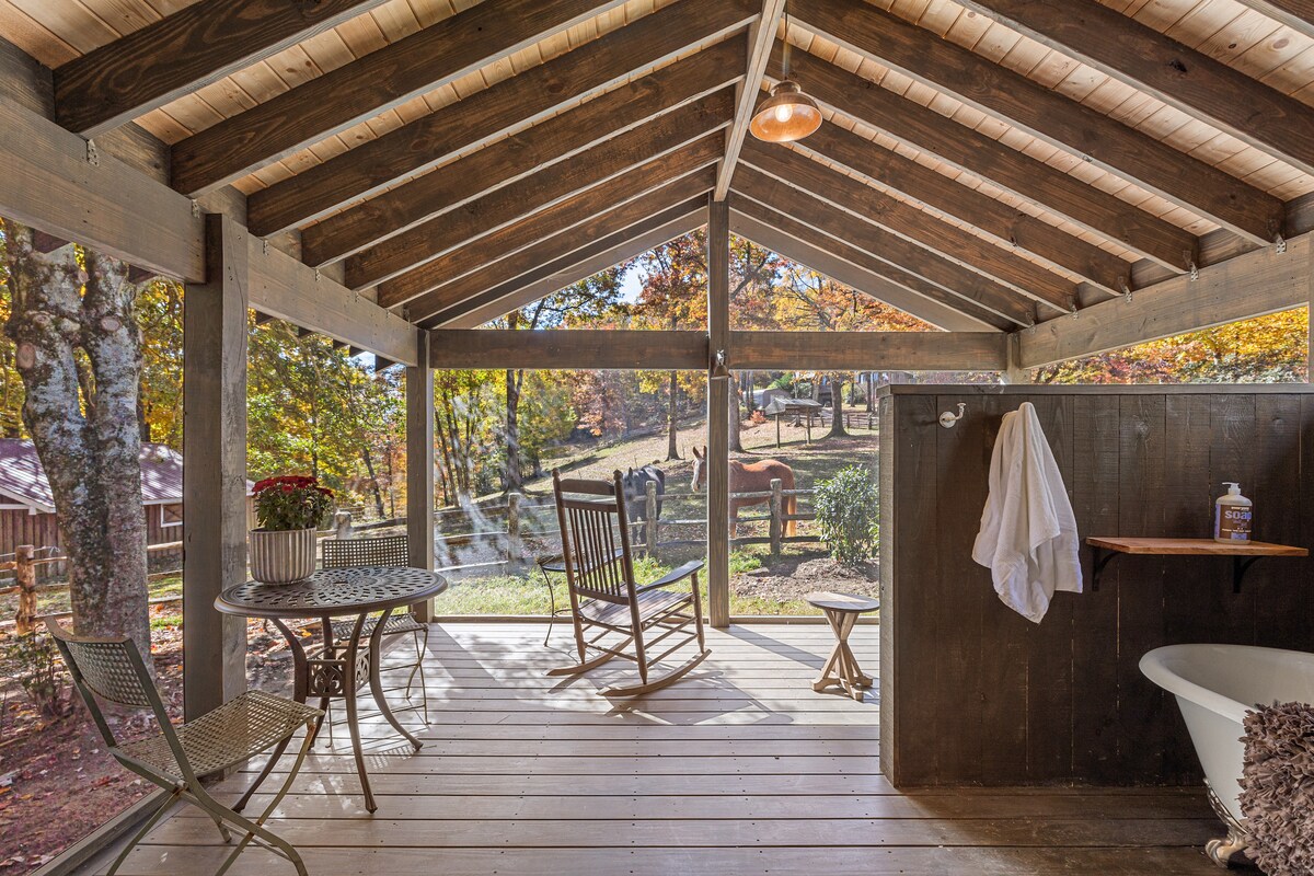 An outdoor patio is showcased with a rustic wooden ceiling and open beams. A comfortable rocking chair and small table create a cozy seating area. The clawfoot tub with a towel hanging nearby is positioned against the privacy panels, overlooking a scenic view of the surrounding nature.