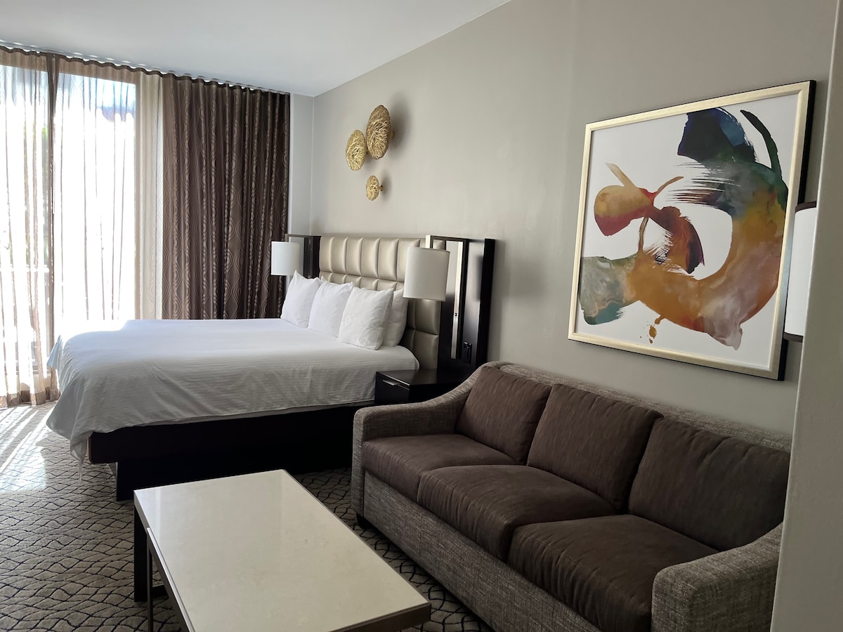 A spacious studio unit is shown, featuring a king size bed with soft white linens. A modern gray sofa is positioned nearby, alongside a coffee table. A large artwork in vibrant colors is displayed on the wall. Natural light enters through expansive window treatments.