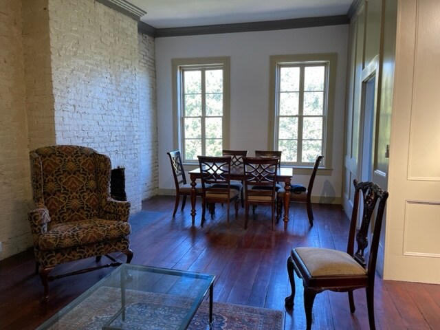 A well-lit living area features hardwood floors and white brick walls. A vintage patterned chair anchors the space beside a glass coffee table. A dining table with six wooden chairs is positioned near the windows, offering views of the outside greenery.