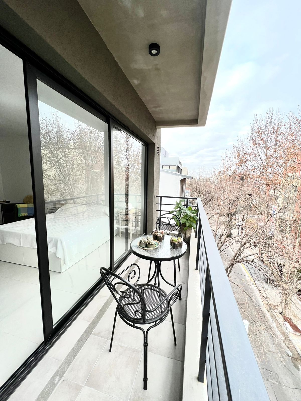 A private balcony features a small round table and two metal chairs, positioned against large glass doors. Outside, bare trees line the street, and the balcony is illuminated with natural light, creating a serene outdoor space.