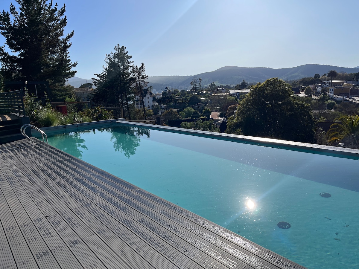 An infinity pool is visible, reflecting sunlight and surrounded by wooden decking. Nearby trees and greenery create a natural border, while hills in the background add to the serene atmosphere. The clear sky enhances the open view from this elevated space.