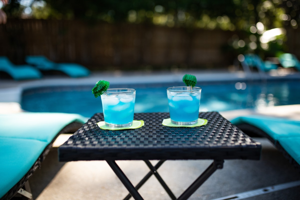 Two refreshing blue cocktails are served on a small, black wicker table beside a sparkling pool. Lounge chairs are positioned nearby, and soft sunlight illuminates the scene, enhancing the inviting outdoor space.