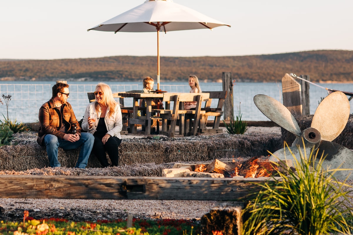 A cozy outdoor seating area is showcased, featuring a wooden table with an umbrella and surrounding chairs. A fire pit with gathered stones provides warmth, bordered by straw bales. Vibrant greenery and a calm water view create a serene backdrop.