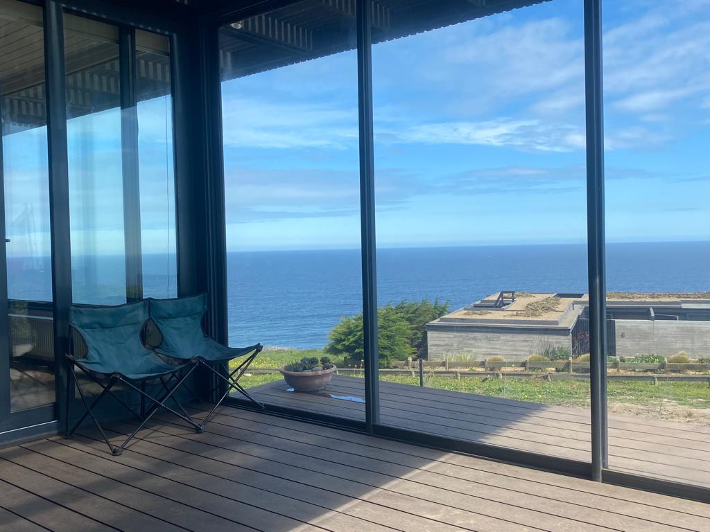 A covered terrace features two blue folding chairs positioned against large glass doors, providing a clear view of the ocean. The expansive sea is visible in the background, with a few coastal structures and greenery lining the horizon under a partly cloudy sky.
