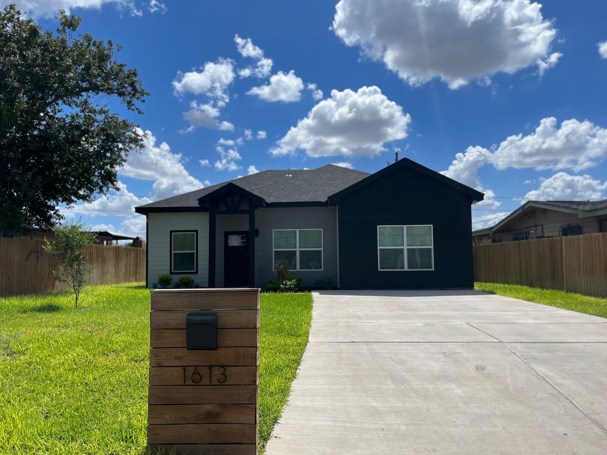 A contemporary one-story house is showcased against a bright blue sky with scattered clouds. The home features a combination of dark and light exterior tones, large front windows, and is set in a spacious yard with a well-maintained lawn. A modern mailbox is visible at the front.