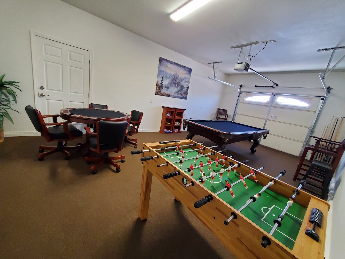A recreational space is shown, featuring a foosball table and a billiards table. A round table with four chairs is present, alongside a wooden cabinet. A garage door and wall-mounted lighting are also visible, contributing to the casual atmosphere.