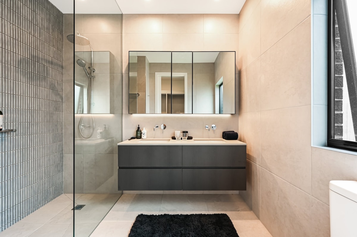 A modern bathroom features a spacious walk-in shower with a glass door and sleek tile walls. A double vanity with a minimalist design is complemented by large mirrors above. Natural light enters through a window, illuminating the neutral-toned tiles and a plush black rug on the floor.