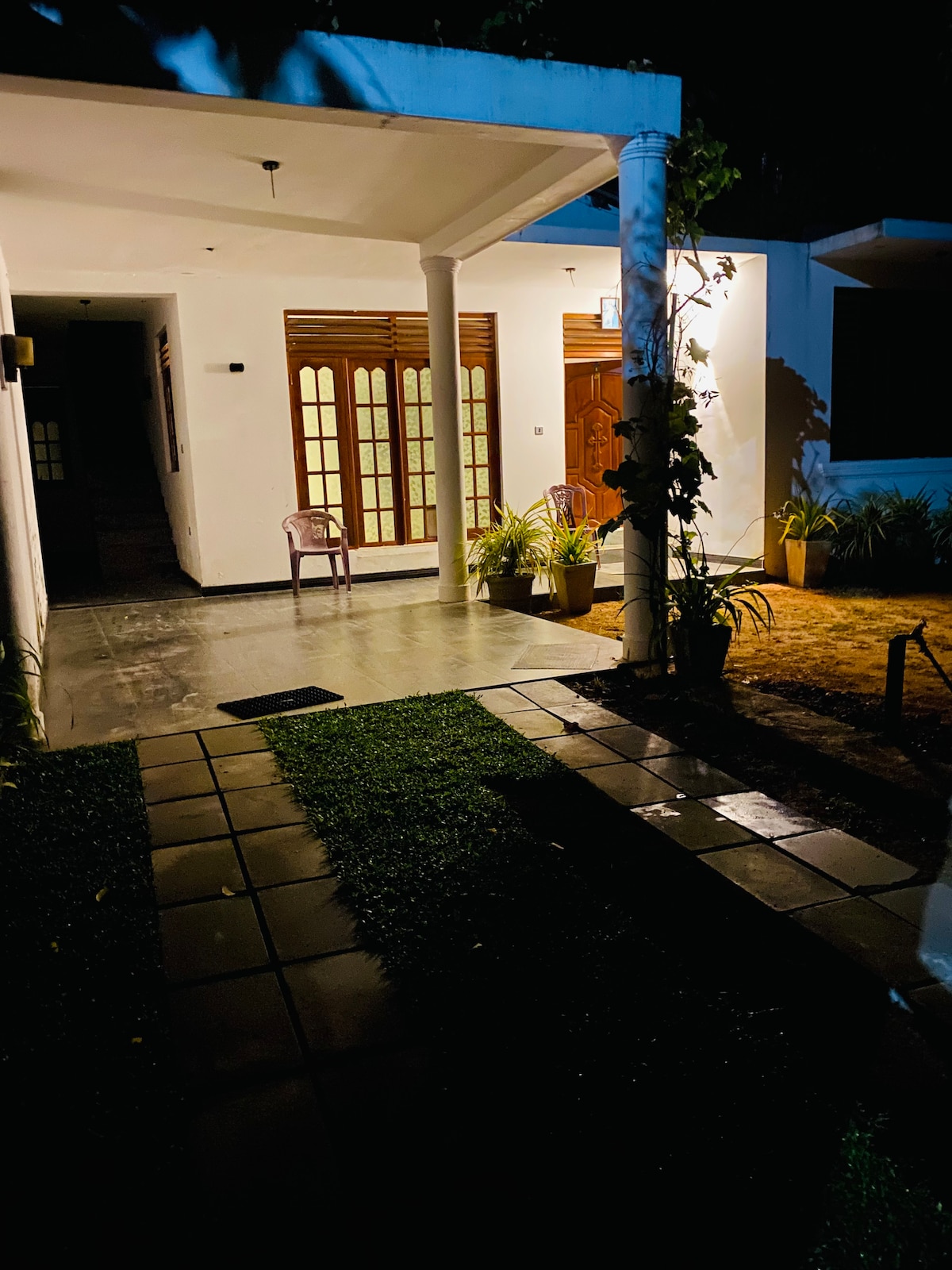 The entrance features a covered porch with two decorative pillars. A stone pathway leads to the front door, surrounded by neatly trimmed grass and potted plants. Soft lighting highlights the architectural details, creating an inviting ambiance at night.