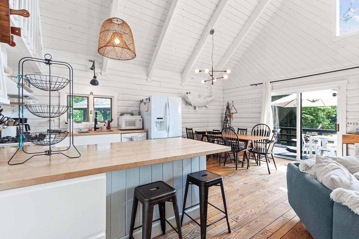 An open kitchen and dining area feature a large wood-topped island with metal stools. Sunlight enters through large windows, illuminating the white walls and exposed wooden beams above. A dining table with black chairs is positioned nearby, along with sliding glass doors leading to an outdoor deck.
