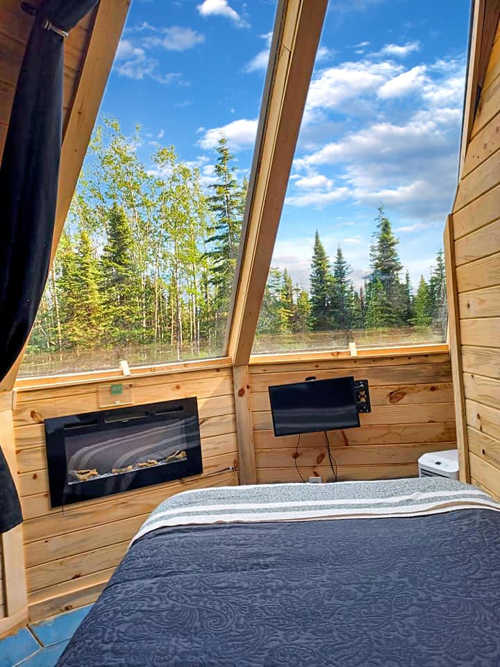 A cozy interior showcases a spacious bed with a dark blanket, positioned near large windows that provide views of the surrounding trees and sky. A wall-mounted TV and electric fireplace are present, enhancing the comfort of the space. Natural light illuminates the wooden walls.