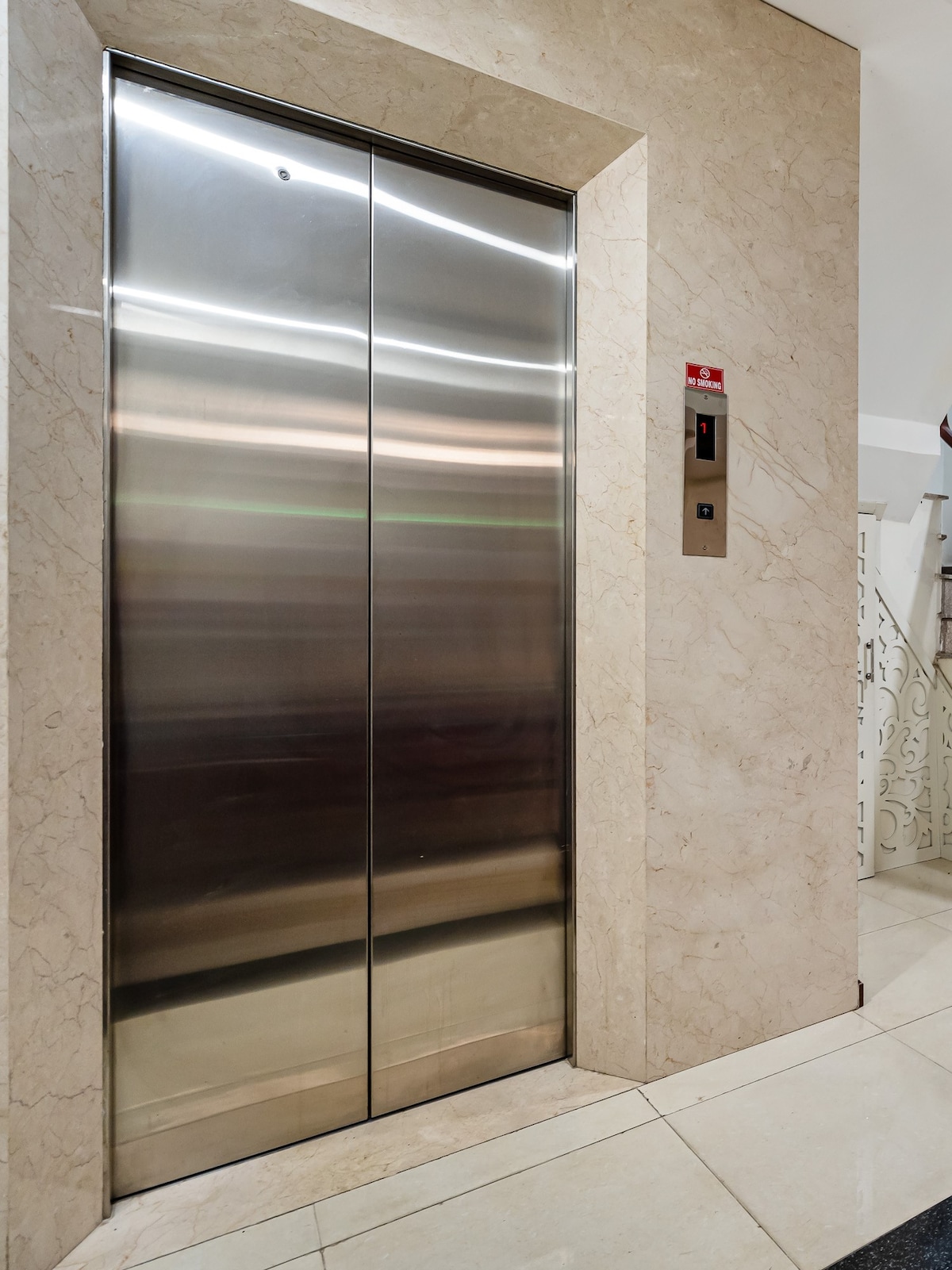 An elevator door is shown against a marbled wall, featuring a sleek stainless steel finish. The control panel is positioned to the side, with buttons for floor selection clearly displayed.