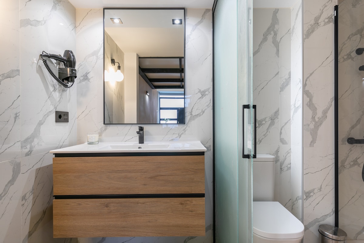 A modern bathroom features sleek marble walls and a sophisticated wooden vanity with dual drawers. A large mirror enhances the space, while a walk-in shower with a glass partition is complemented by a contemporary black shower fixture and a minimalistic toilet.