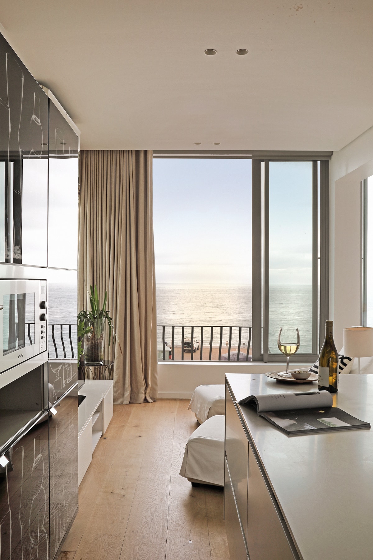 A modern kitchen area is illuminated by natural light from large windows overlooking the ocean. A small table is set with a wine glass and a bottle, while a decorative plant adds a touch of greenery. Soft curtains frame the view, enhancing the serene atmosphere.