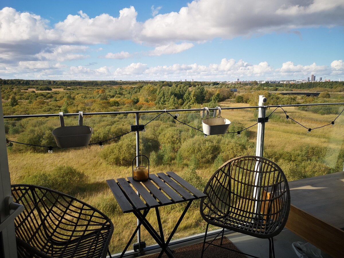 A spacious balcony is showcased with two woven chairs and a small wooden table. The view encompasses lush greenery and open fields, extending towards a distant city skyline under a partly cloudy sky. Soft lighting adds a serene touch to the outdoor space.