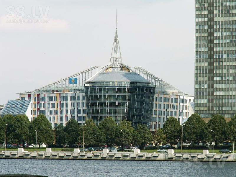 A modern building is depicted, featuring a distinctive angular rooftop spire. The structure is characterized by a large glass facade, reflecting the surrounding environment. Trees are visible along the waterfront, with the river seen in the foreground, enhancing the urban landscape.