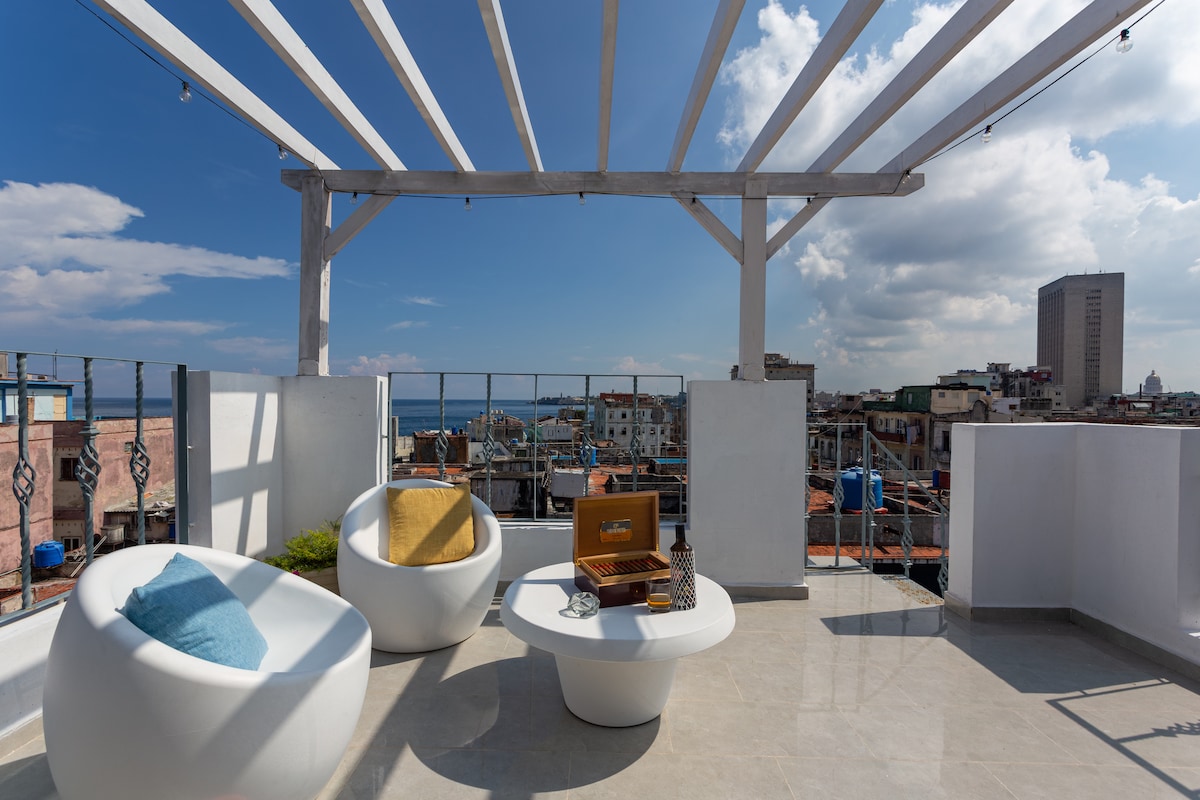 A spacious rooftop terrace features modern white furniture, including a round table and comfortable chairs. The area is brightly lit by natural sunlight, with strings of lights overhead and a panoramic view of the city and sea in the background.
