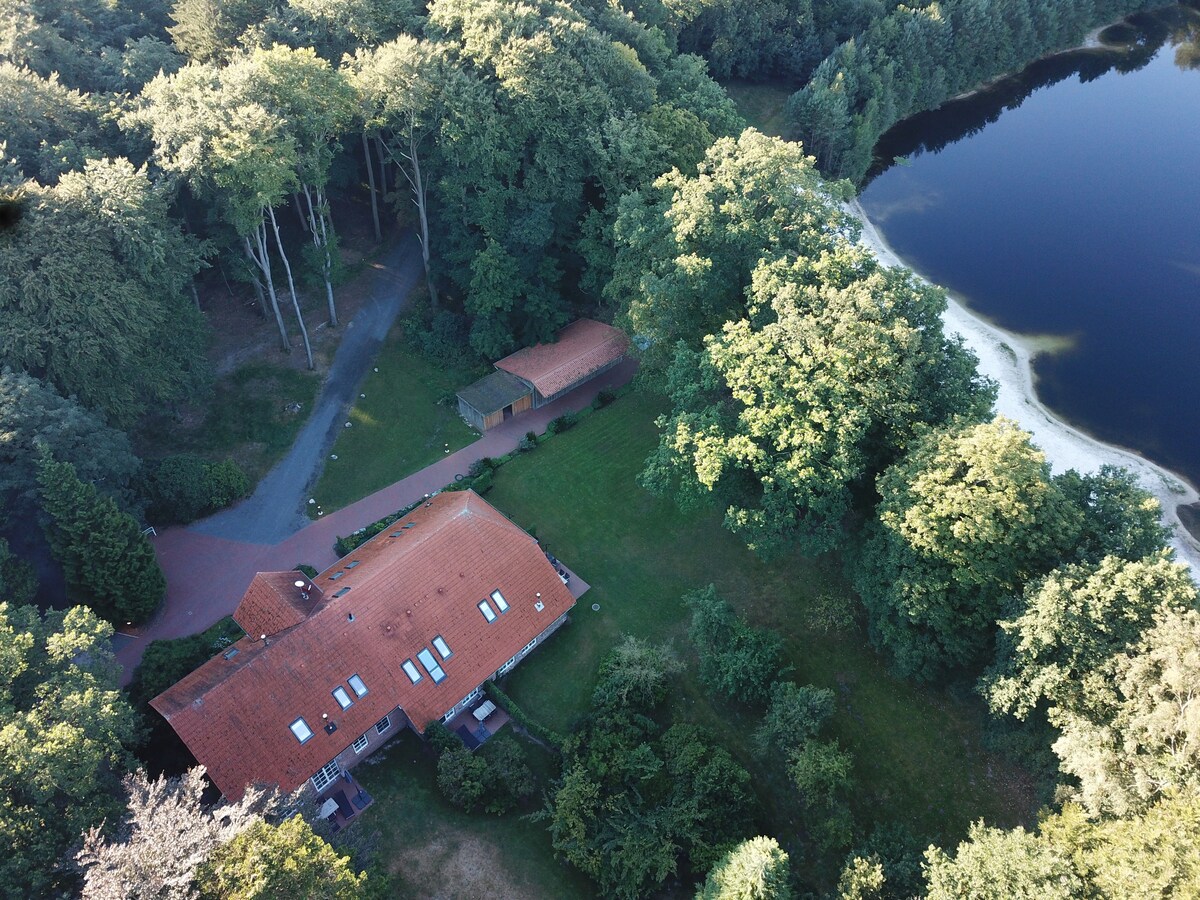 An aerial view showcases a sprawling building surrounded by lush trees and a private lake. The main structure features a red-tiled roof and multiple skylights, while pathways lead to the water's edge. The natural environment enhances the serene setting.
