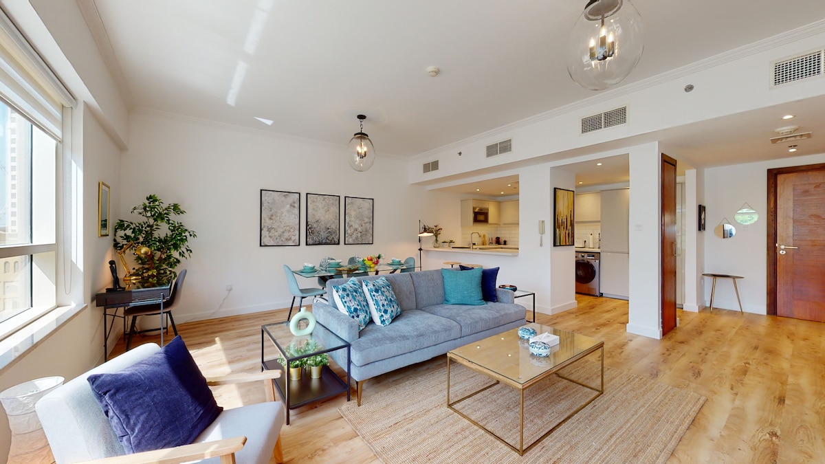 A modern living area features a spacious gray sofa adorned with colorful cushions. A glass coffee table sits on a light wood floor, surrounded by plants and artwork. An open kitchen and dining area are visible in the background, enhancing the overall bright and airy ambiance.