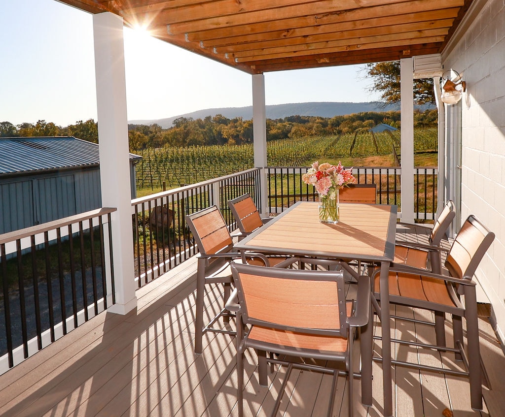 An outdoor deck features a spacious dining table surrounded by six chairs. A vase of flowers is placed in the center. The backdrop showcases rolling vineyards and a mountain range under a warm, golden sunlight.