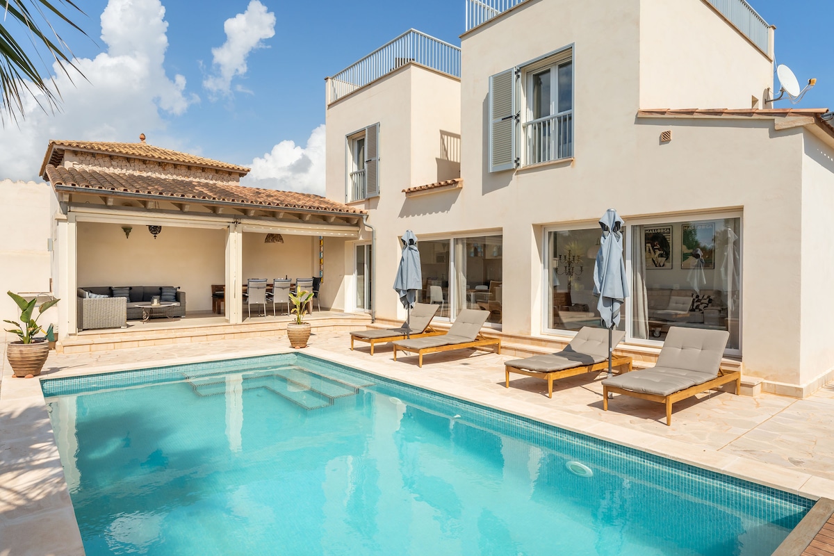 A spacious pool area is featured in the foreground, with a clear blue pool reflecting the sky. Surrounding the pool are several lounge chairs, and a covered seating area is visible in the back, providing shade and comfort.